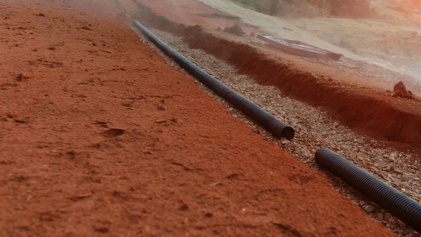 Open French-drain trench mid-install with washed gravel and perforated pipe.