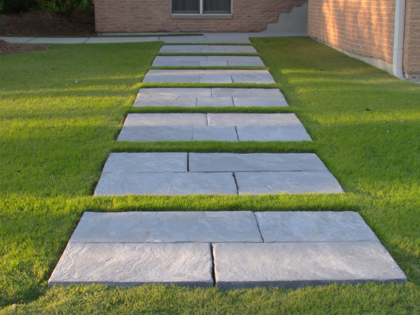 Square 24 inch bluestone stepping stones spaced across a Bermuda grass side yard at a single-story brick Texas home