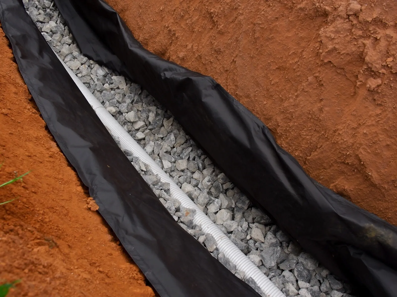 Open French drain trench with white 4 inch perforated pipe surrounded by angular crushed gray lateral drainage rock and landscape fabric
