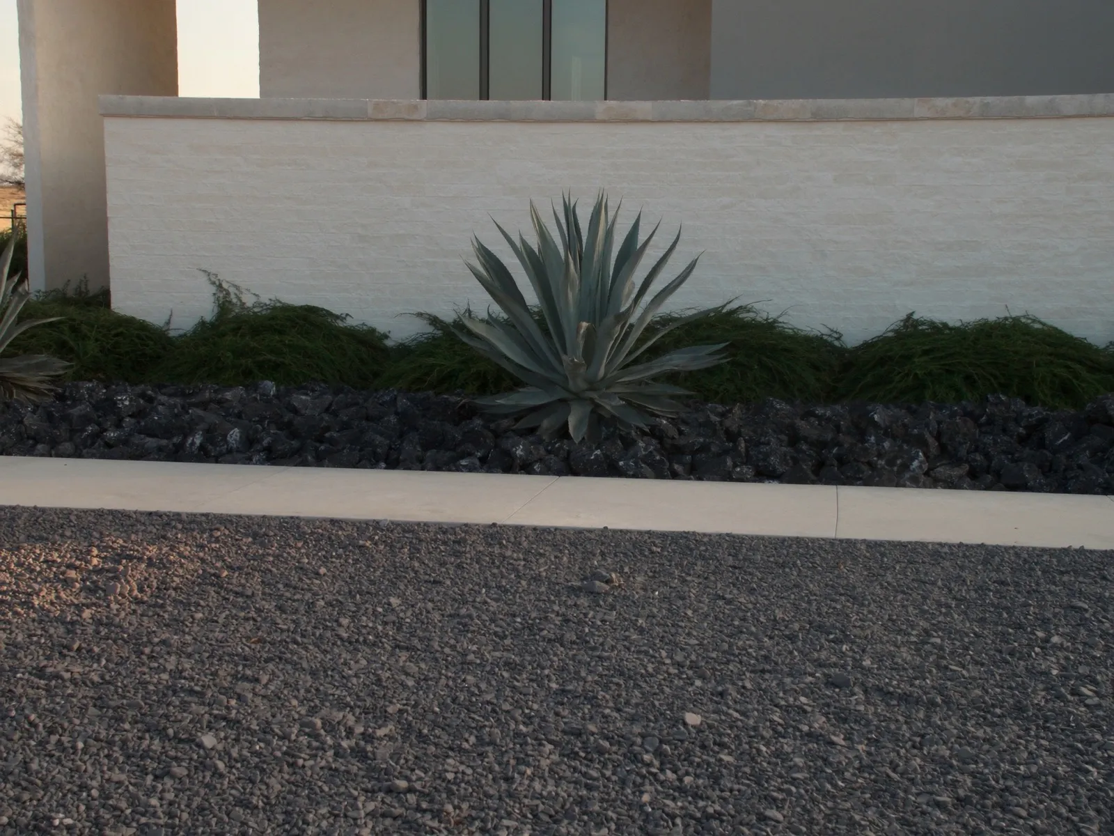 Modern xeriscape bed of black decorative rock with a single architectural agave in front of a limestone and stucco Texas home