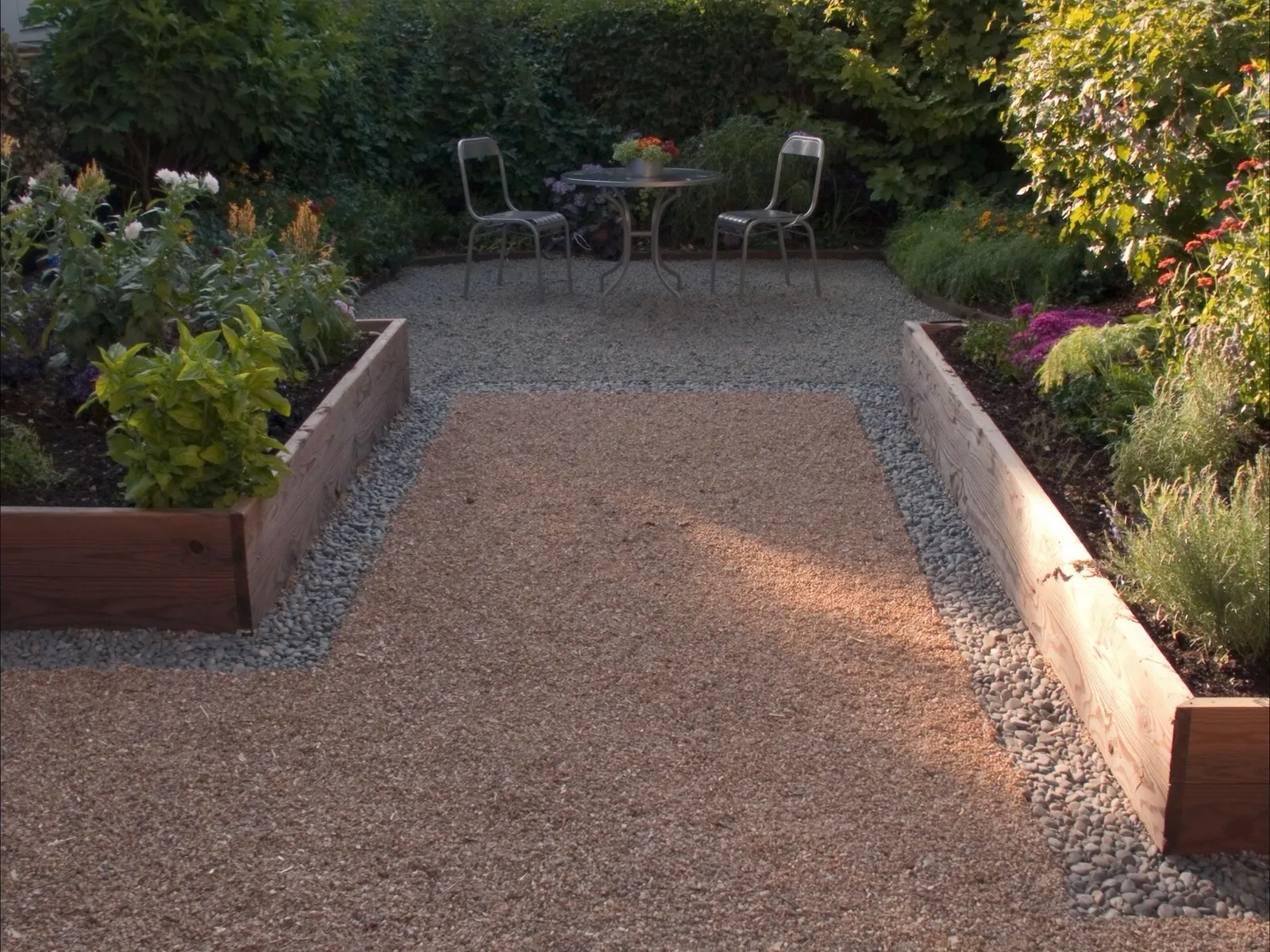 Pea gravel garden path between two wooden raised beds leading to a small pea gravel patio with a bistro table at golden hour