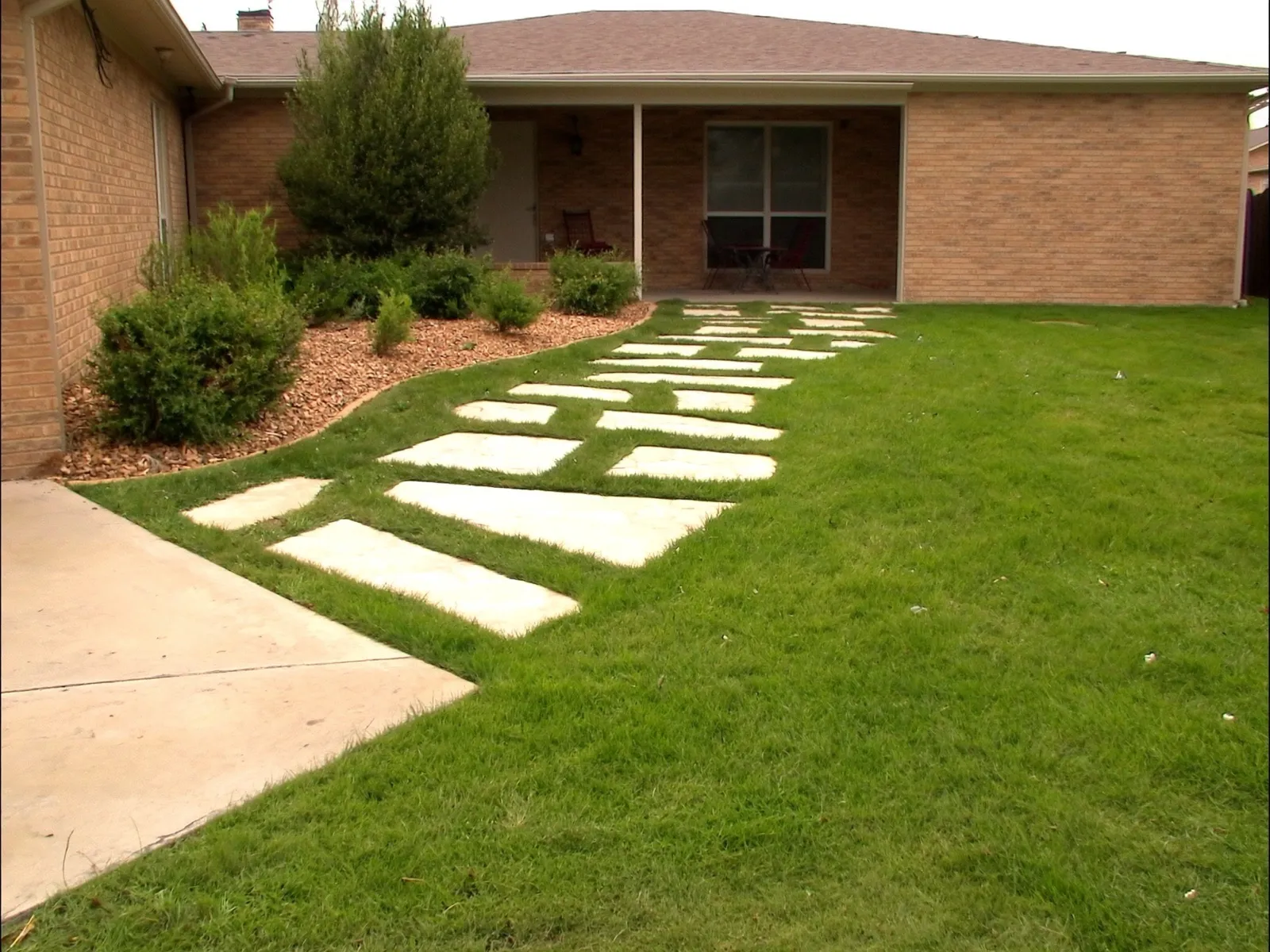 Cream Texas flagstone walkway across a Bermuda lawn to the front porch of a single-story tan-brick Texas home
